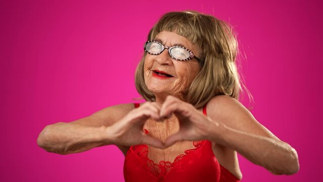 Funny portrait of elderly old woman toothless, giving heart gesture with hands isolated on pink background wearing wig, glasses, negligee