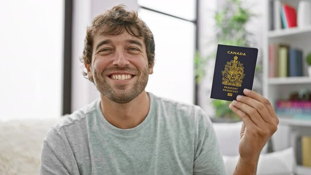 Happy young man, smiling and sitting at home on sofa, holding his canadian passport, ready for vacation!