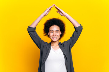 Happy curly haired african american or brazilian girl, making roof of hands above head, looking at camera with toothy smile, feeling herself in safety,dreaming about housing,isolated orange background