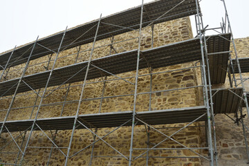 Metal scaffolding set up against a stone wall, likely for construction or restoration work. The scaffolding has multiple levels and frames the rough textured wall behind it.
