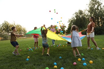 Group of children and teachers playing with rainbow playground parachute on green grass. Summer...