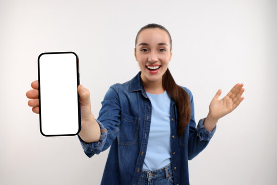 Surprised Young Woman Showing Smartphone In Hand On White Background, Selective Focus. Mockup For Design