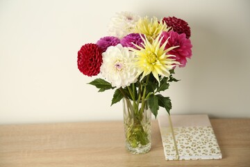 Bouquet of beautiful Dahlia flowers in vase and notebook on wooden table near white wall