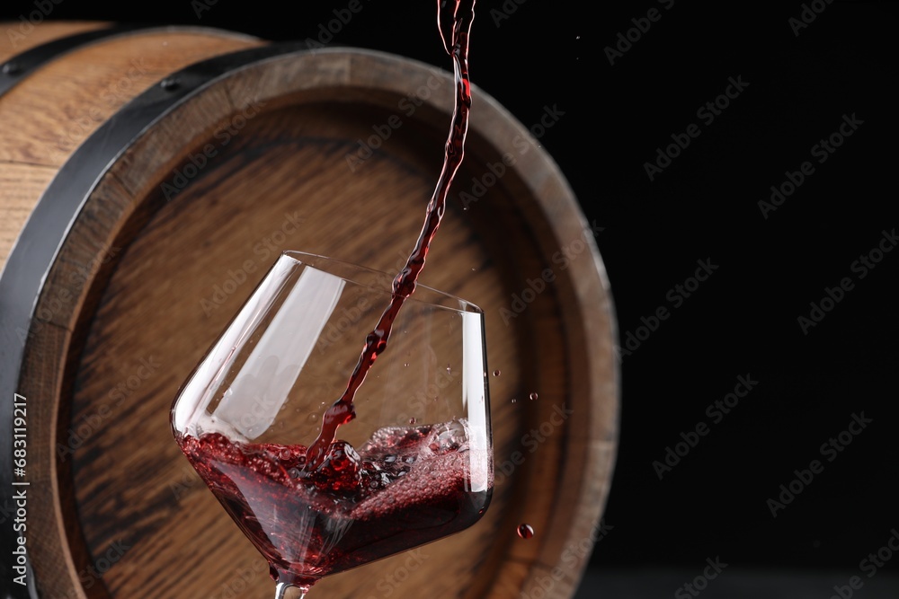 Wall mural Pouring delicious wine into glass near wooden barrel against black background, closeup. Space for text