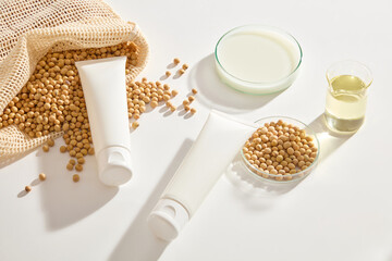 Blank label tubes in white color displayed with a mesh bag of soybeans and glassware filled with milk and oil. Soybean is associated with reducing the visible effects of skin aging