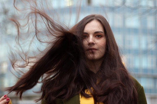 Street photo. Beautiful young white childfree brunette woman against background of glass wall of building with windows. Wind in loose hair, covering her face. Concept of freedom, beauty, naturalness
