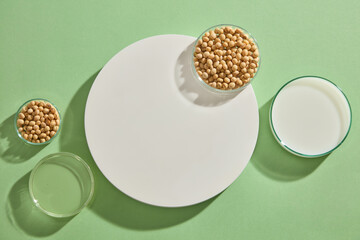 Flat lay of some glass petri dishes of soybean seeds, milk and oil displayed on green background with a round podium. Mock-up for exhibitions or presentation of cosmetic products or packaging