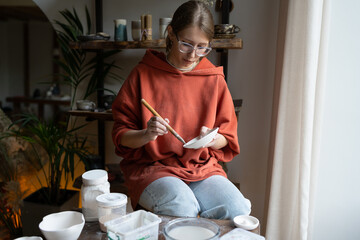 Creative pleased woman artisan applying protective solution with brush on earthenware preparing...