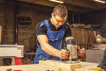 A young  man wearing safety goggles carpenter builder pre-works the edges of the wooden board with...