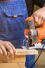 Close up of experienced carpenter in work clothes and small buiness owner  carpenter saw and processes the edges of a wooden bar with a jig saw  in a workshop