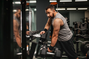 A bearded male bodybuilder takes a dumbbell in his hand and demonstrates the triceps. Work on your body in the gym.