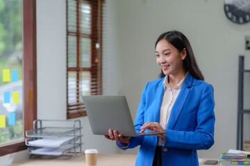 Attractive smiling Asian businesswoman standing holding laptop computer working in office.