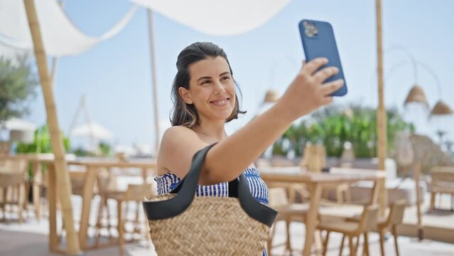 Young hispanic woman taking a selfie picture standing at sunny restaurant terrace