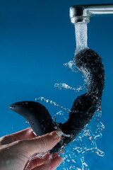 A woman washes a black prostate stimulator under running water on a blue background. Sex toy...