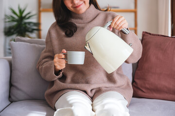 Closeup image of a young woman pouring hot water from electric kettle in to a cup at home