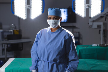 Portrait of biracial female surgeon wearing surgical gown and face mask in operating theatre