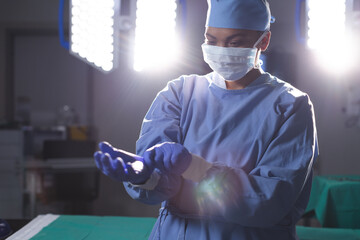 Biracial female surgeon wearing surgical gown and medical gloves in operating theatre - Powered by Adobe