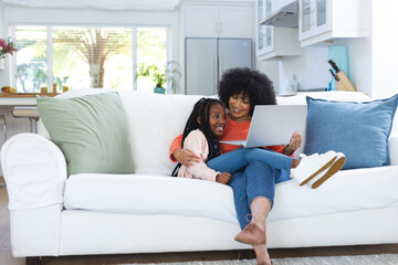 Happy african american mother and daughter embracing on couch and using tablet at home, copy space