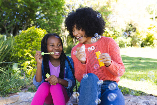 Happy african american mother and daughter sitting and blowing bubbles in sunny garden, copy space
