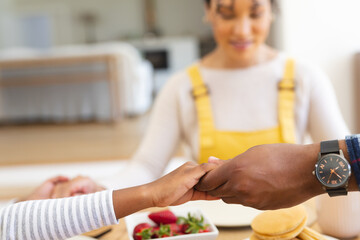 African american family holding hands and praying at table in dining room at home, copy space