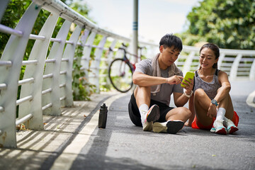 young asian couple sitting on ground using cellphone outdoors in park - obrazy, fototapety, plakaty