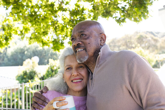 Happy diverse senior couple holding cups of coffee and embracing on sunny terrace