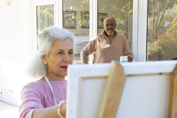 Diverse senior woman painting on canvas and senior man drinking coffee on sunny terrace