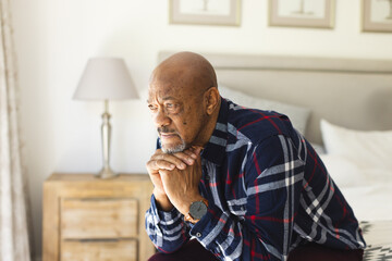 Thoughtful african american senior man sitting on bed in sunny bedroom