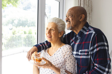 Diverse senior couple drinking tea, embracing and looking out window at sunny home