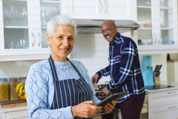 Happy diverse senior couple preparing meal using tablet in kitchen