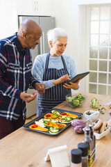 Diverse senior couple preparing meal using tablet in kitchen