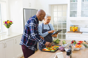 Diverse senior couple preparing meal using tablet in kitchen