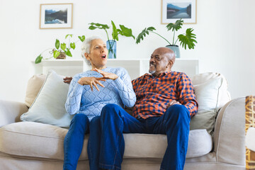 Happy diverse senior couple sitting on sofa, talking and embracing in sunny living room