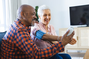 Happy diverse senior man testing blood pressure of senior woman in sunny living room
