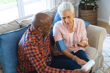 Diverse senior man testing blood pressure of senior woman in sunny living room