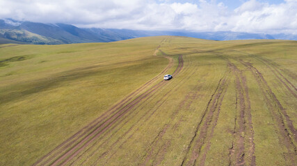 A white SUV is driving in the mountains through the fields. Aerial view from a drone of huge white clouds, snowy mountain peaks and green fields. Dirty off-road with puddles. Splashes from the wheels