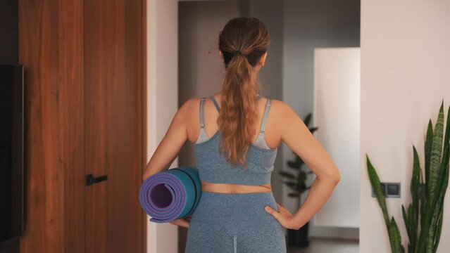 young happy woman going to do sports at home back rear view, holds karimat mat in hands, sportswear suit blue top leggings. modern room interior. aerobics sport fit yoga workout domestic fitness 4k