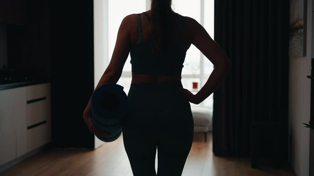 young happy woman going to do sports at home by window back rear view dark silhouette, holds karimat mat in hands, sportswear suit . modern room interior. sport fit yoga workout domestic fitness 4k