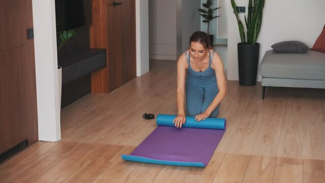 young happy woman holds karimat in hands rolls up folds mat cleans up after training at home. sportswear suit blue top leggings. modern room trendy interior. aerobics sport fit yoga workout fitness 4k