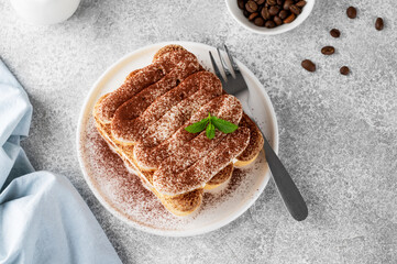 Tradition italian layered dessert tiramisu with mascarpone cream and biscuits on a white plate with cup of coffee on a gray concrete background.