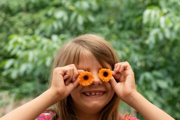  whimsical young girl with delighted grin uses orange calendula flowers to mimic eyeglasses, covering her eyes, makeshift floral spectacles, embodying spirit of playfulness against garden backdrop