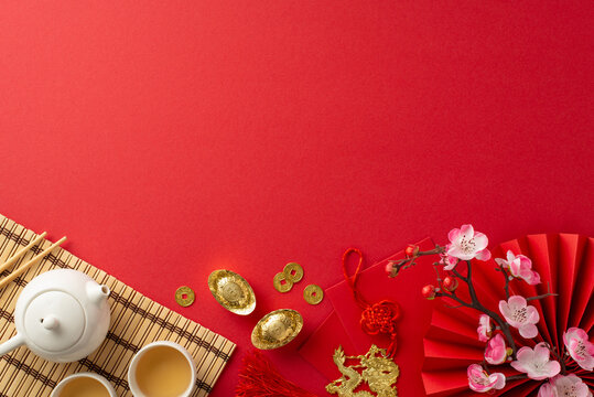 Traditional Chinese New Year Table: top view tea ceremony set, bamboo placemat, coins, red envelopes, dragon charm hanging, sakura and more on red backdrop. Create festive atmosphere for your promo
