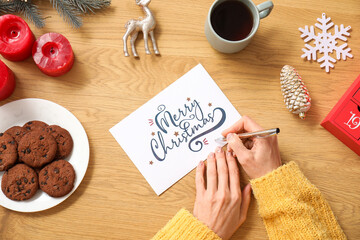 Mature woman writing greeting card for Christmas celebration on wooden table, top view