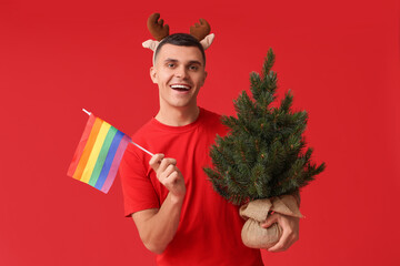 Young man in reindeer horns with LGBT flag and Christmas tree on red background