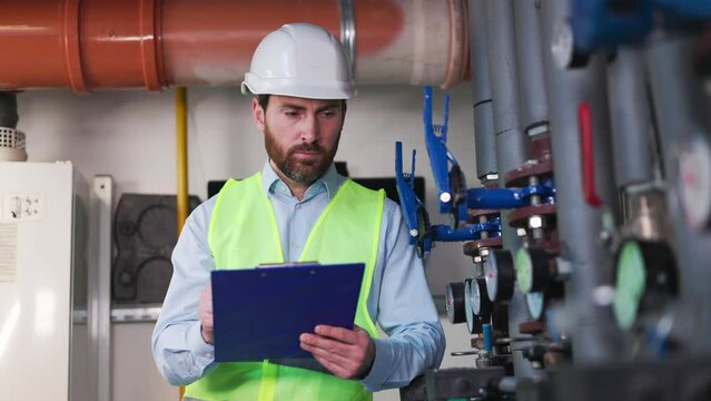 Business manager, mechanic, engineer, plumber holds a tablet and makes notes, recording water meter readings. The factory worker analyzes data, located in the boiler room of a commercial building.