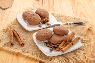Fototapeta na wymiar Chocolate cookies, cinnamon, star anise on ceramic saucer with cup and jute napkin on wooden table.