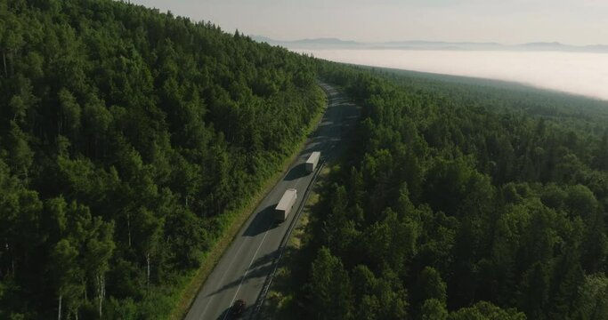 Two Semi Trucks with white trailer and cab driving, traveling alone on dense forest mountain winding road, highway perspective view follow vehicle aerial footage. Freeway trucks traffic