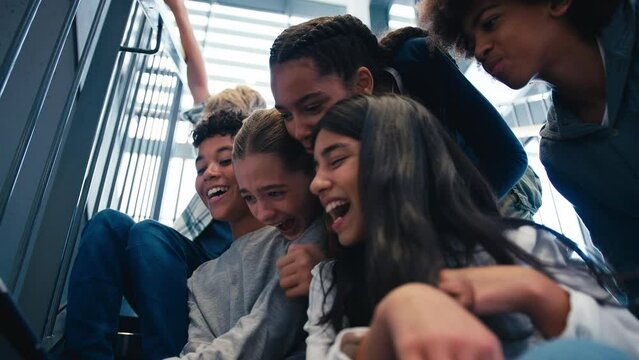 Multi-cultural group of male and female secondary or high school pupils inside school building on stairs pulling silly faces and posing for selfie on digital tablet - shot in slow motion