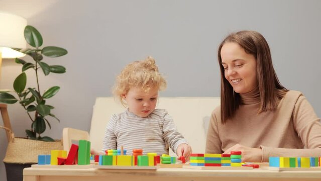 Shape sorter challenge. Playful motor activities. Wood block stacking. Geometric toy exploration. Cute young mother playing with her toddler baby daughter in home interior.