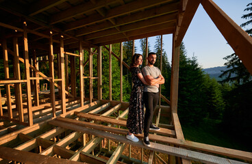 Naklejka na ściany i meble Man and woman inspecting future wooden frame dwelling nestled in the mountains near forest. Youthful couple at construction site in early morning. Concept of contemporary ecological construction.